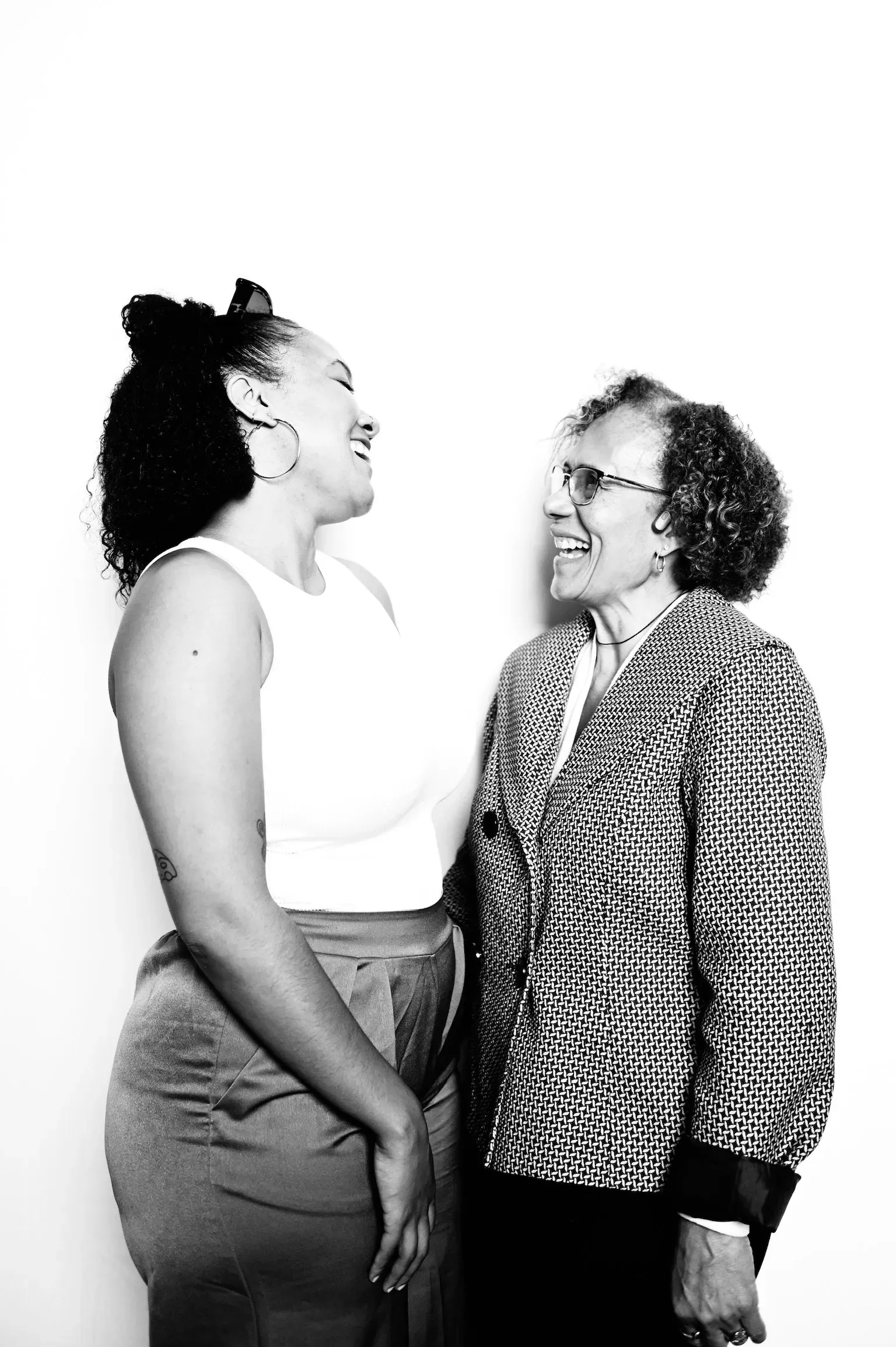 Two women facing each other, smiling, in patterned and white tops against a plain background.