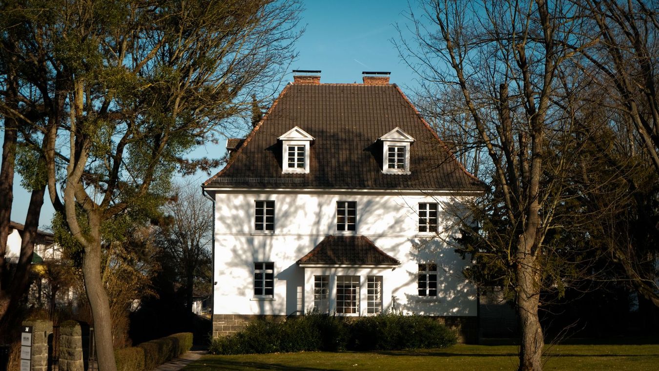 White two-story house with a dark roof and dormer windows, framed by trees on a sunny day