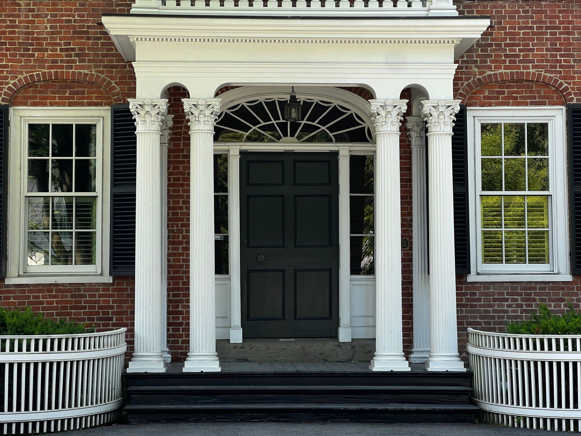 Brick building entrance with white columns, black door, and two windows with shutters.