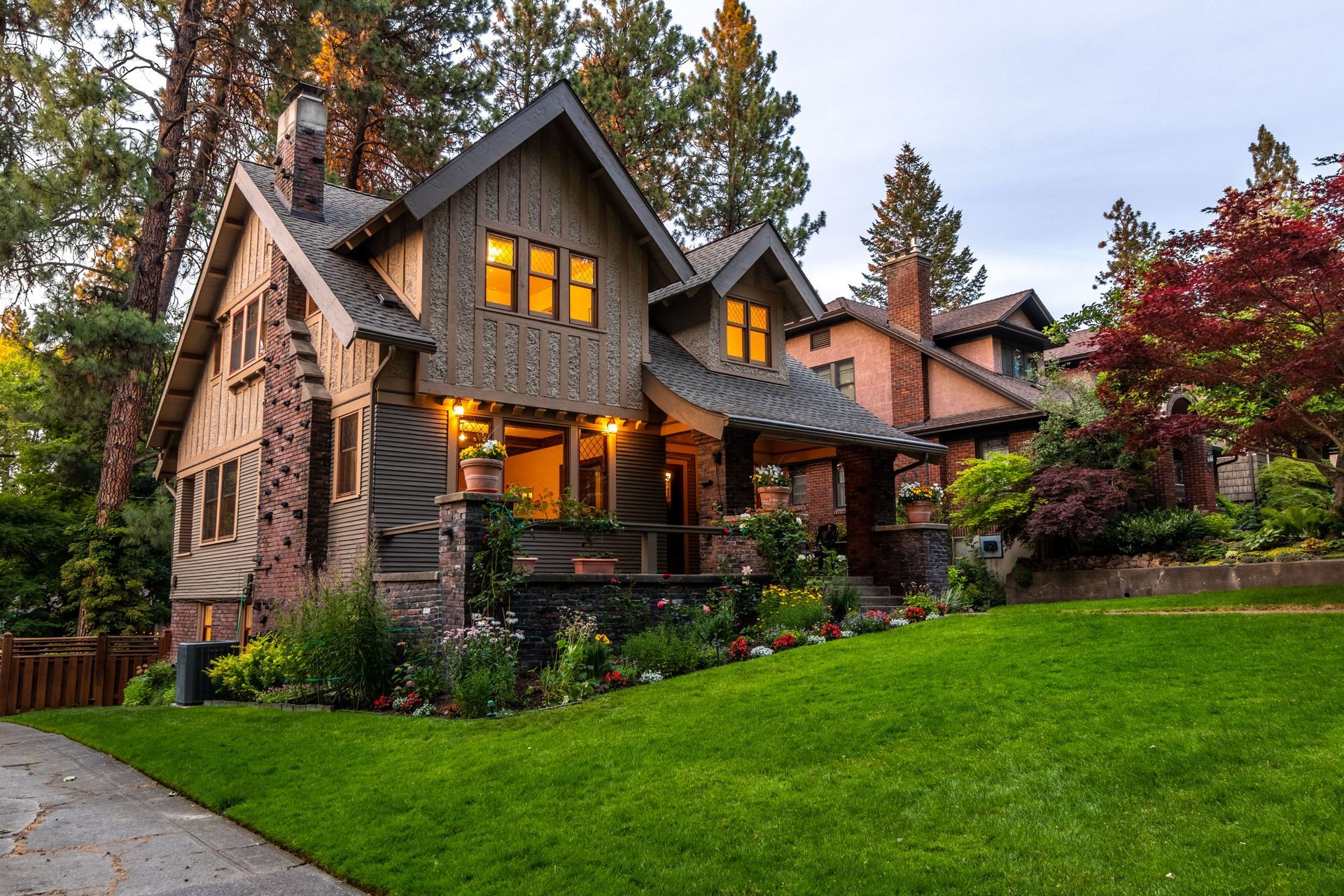 Stone house with warm lit windows, a front porch, and a green lawn surrounded by tall trees at dusk