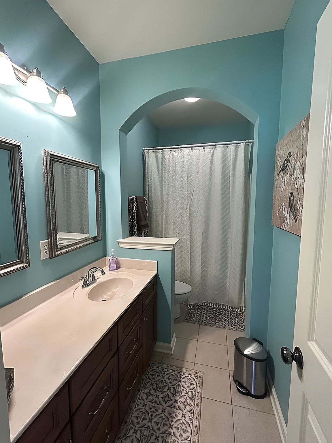 Narrow turquoise bathroom with vanity, mirrors, shower curtain, and patterned floor tile.
