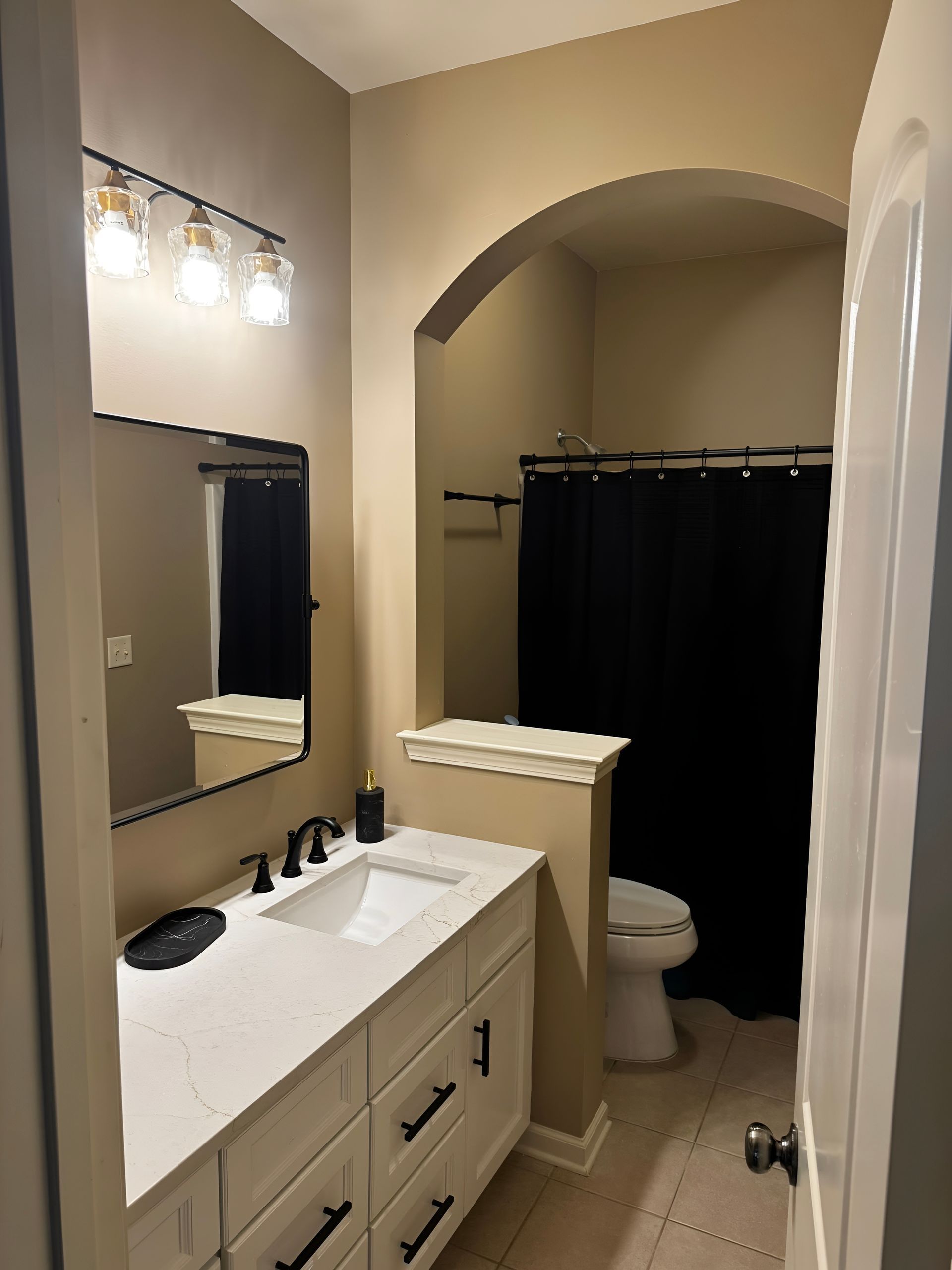 Small bathroom with white vanity, black fixtures, mirror, and black shower curtain in a beige alcove.