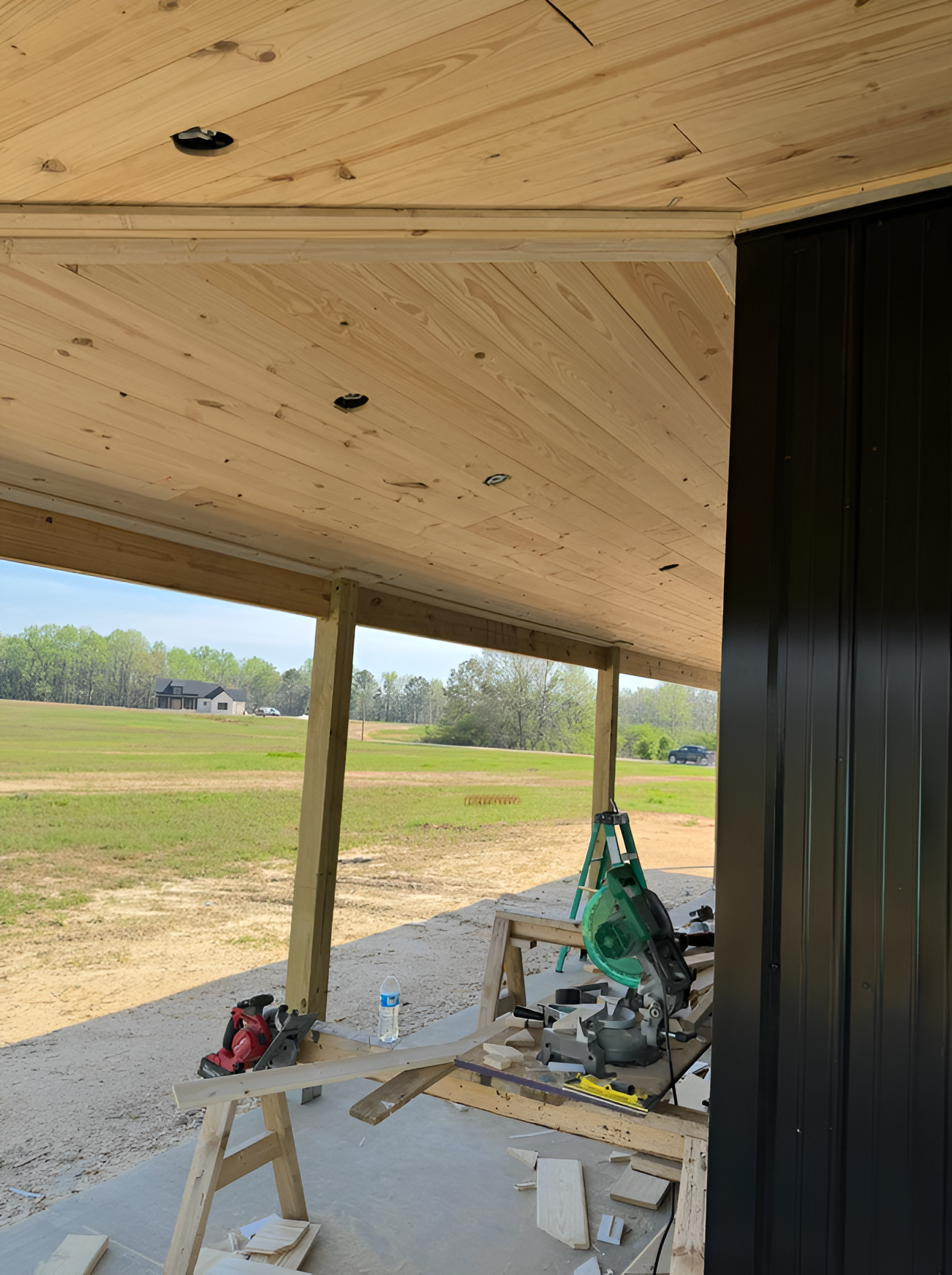 Covered outdoor porch under construction with wood ceiling, posts, tools, and open field beyond