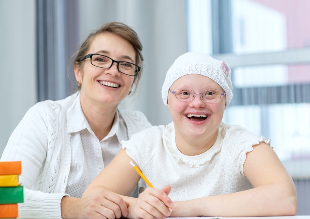 A Woman and a Girl With Down Syndrome Are Sitting at a Table — Adaptive Care Pty Ltd In Tarneit, VIC