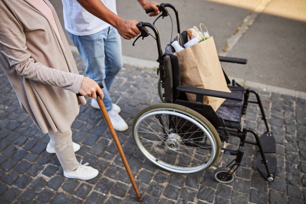 An Aged Woman Leaning on A Walking Stick and A Man by Her Side Pushing a Wheelchair — Adaptive Care Pty Ltd In Tarneit, VIC
