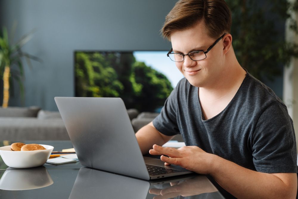 A Man With Glasses Smiles While Using a Laptop — Adaptive Care Pty Ltd In Sunshine, VIC