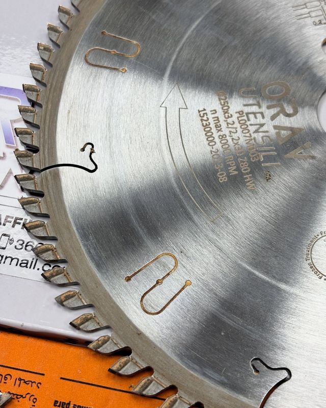 Primo piano di una lama di sega circolare argentata con marcature incise su una superficie arancione.