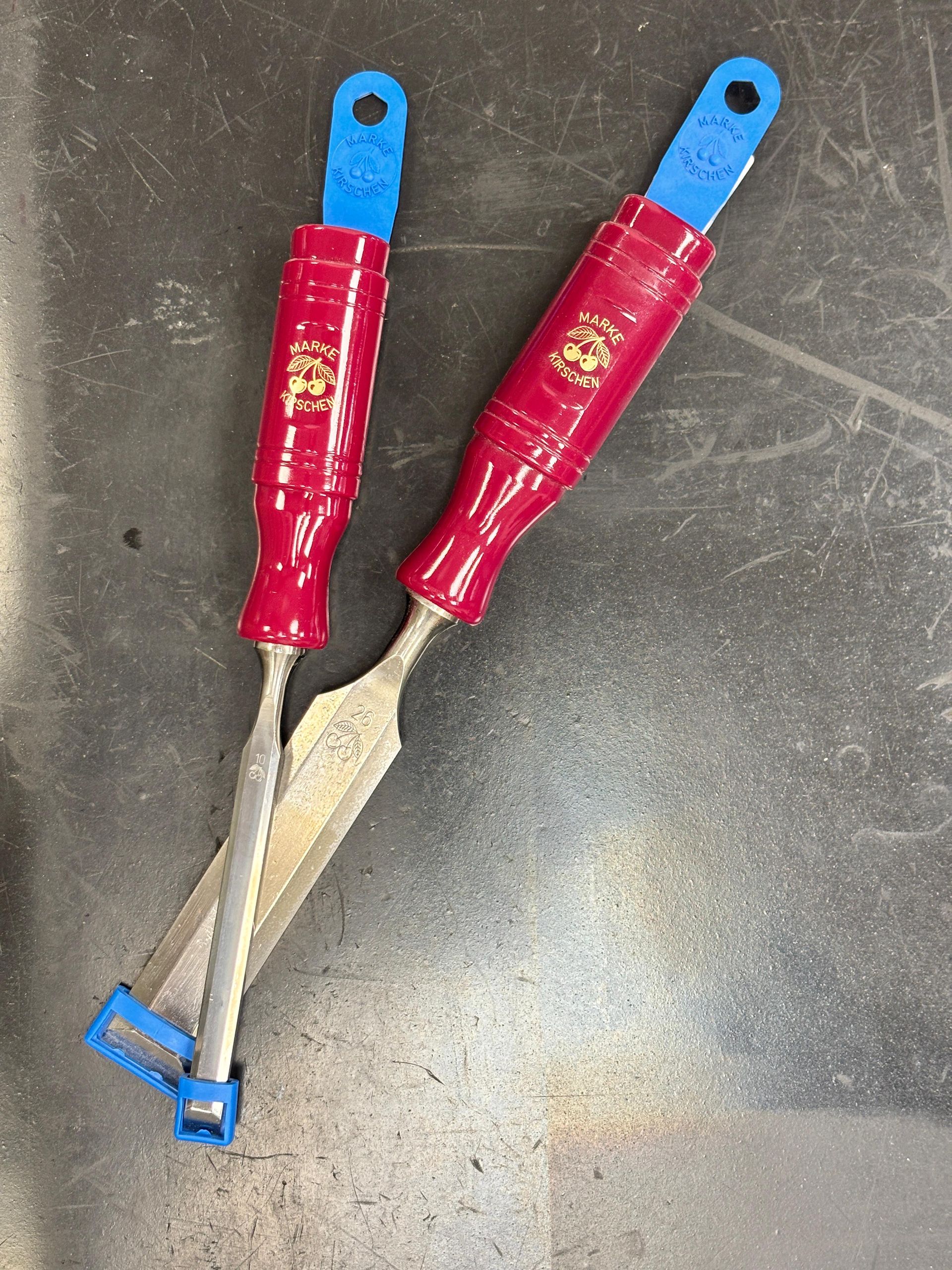 Due scalpelli di metallo con manico rosso e anelli blu per appenderli, appoggiati su un tavolo scuro.