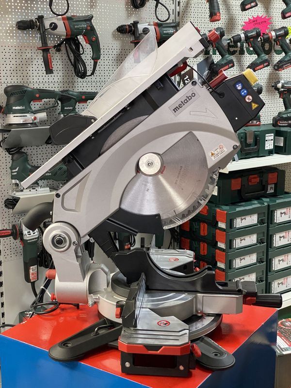 Una sega troncatrice grigia su un banco da lavoro arancione in un'officina, con trapani e contenitori per attrezzi sullo sfondo.