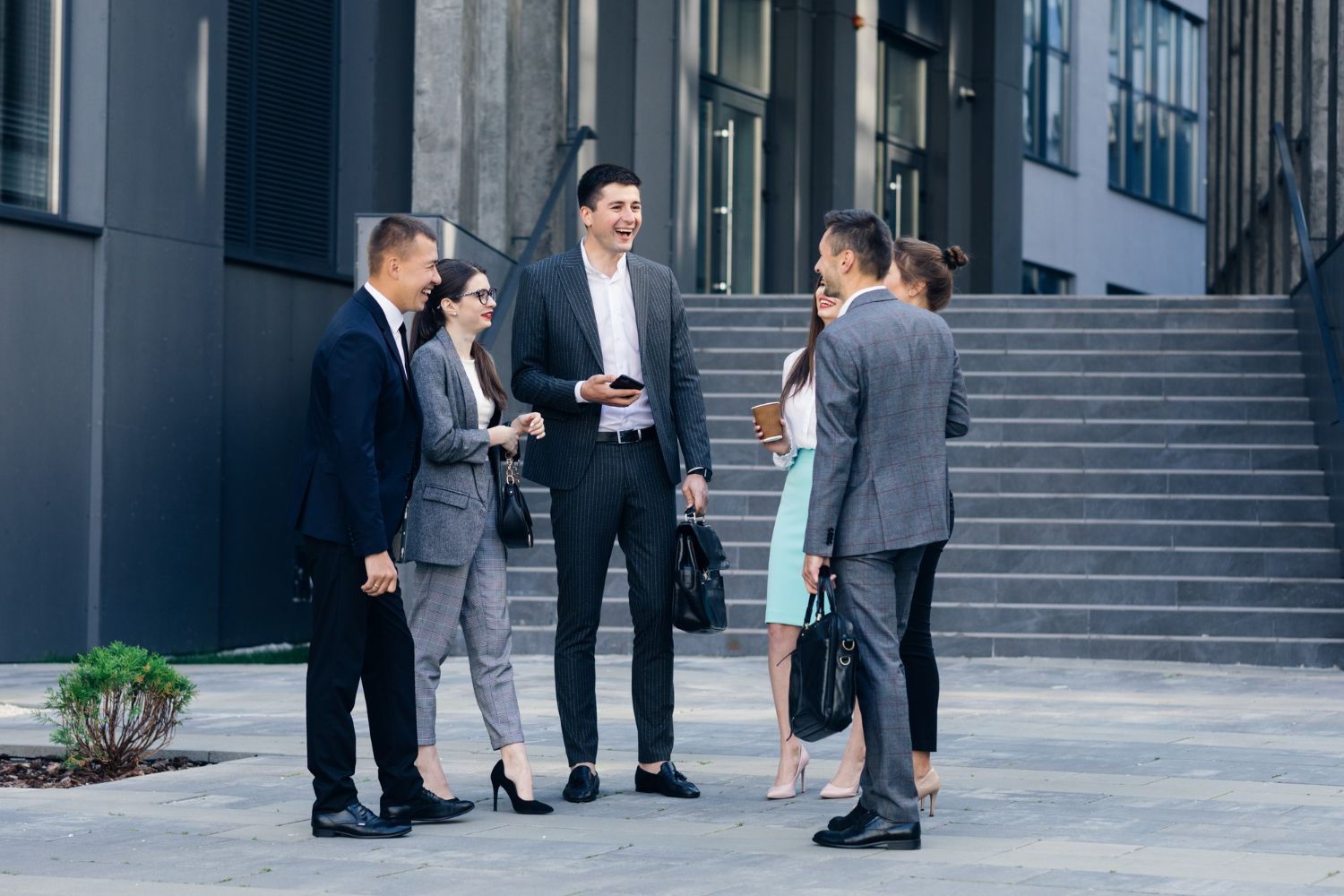 A group of people in business attire stand and talk on a stone plaza in front of a modern office building.