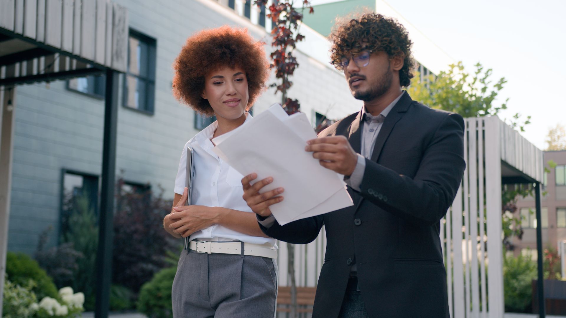 A professional pair walking outside, reviewing documents together near a modern office building.