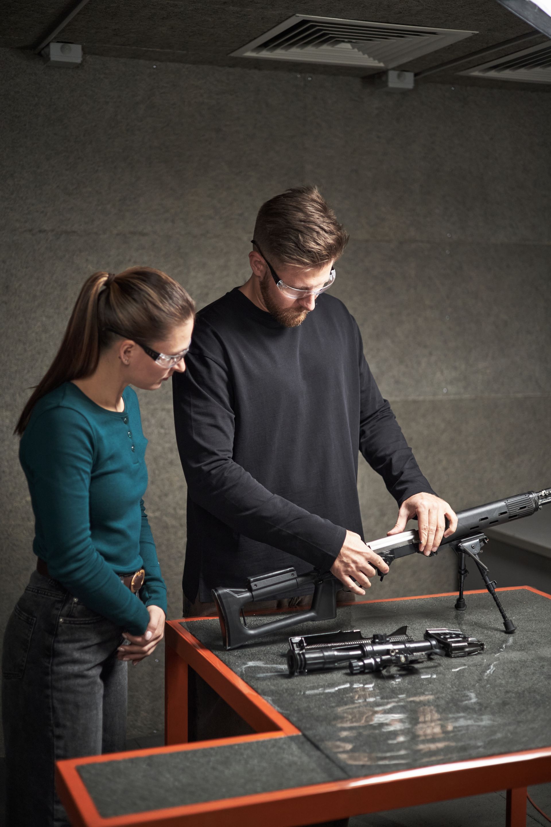 A person in a black shirt teaches another person in a green shirt how to assemble a rifle on a shooting range table.