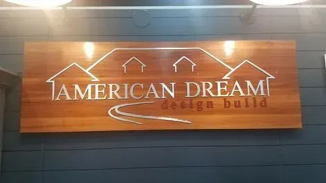 Wooden “American Dream design build” sign with house outline, mounted on a dark wall.