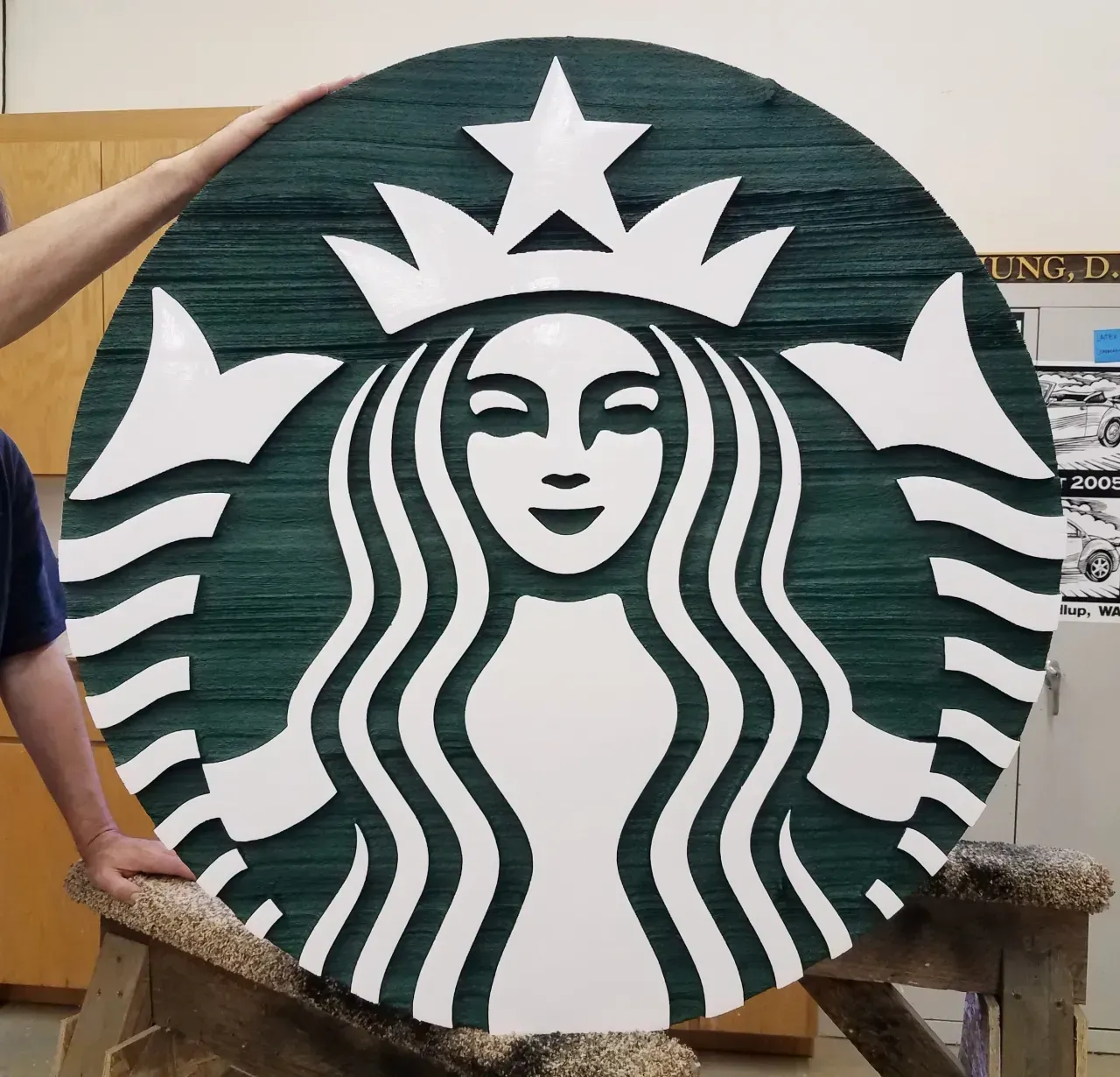 Green Starbucks logo sign on a round wooden disc, held outdoors by a person.