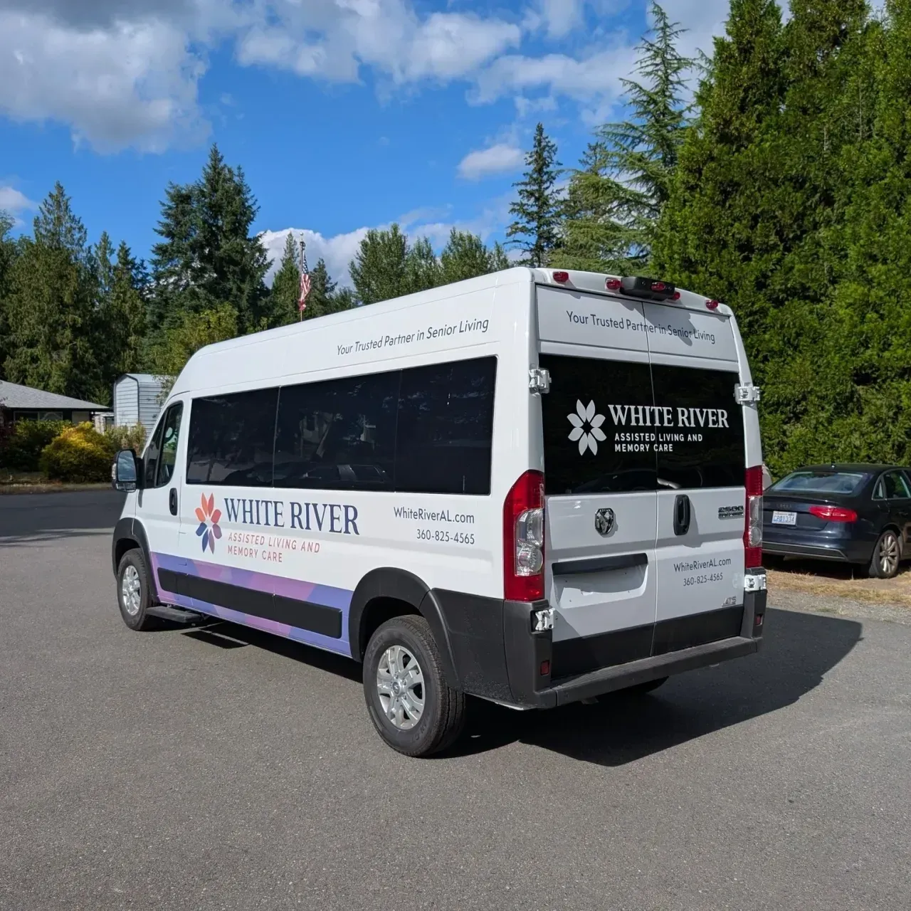 White River transit van parked on a road, white with blue and purple stripes, trees and blue sky behind it