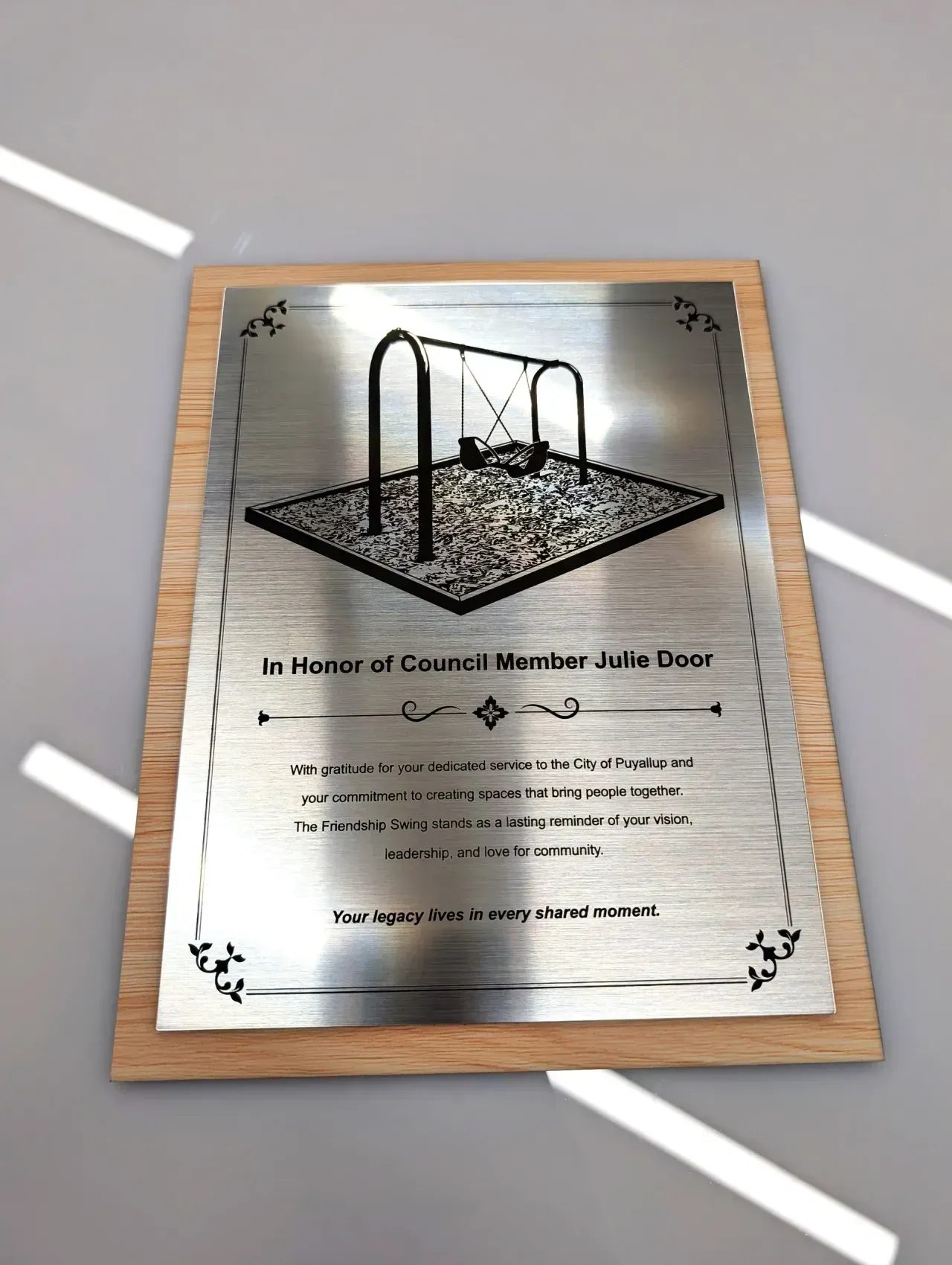 Framed plaque with a black-and-white playground swing illustration and memorial text on a light wood table