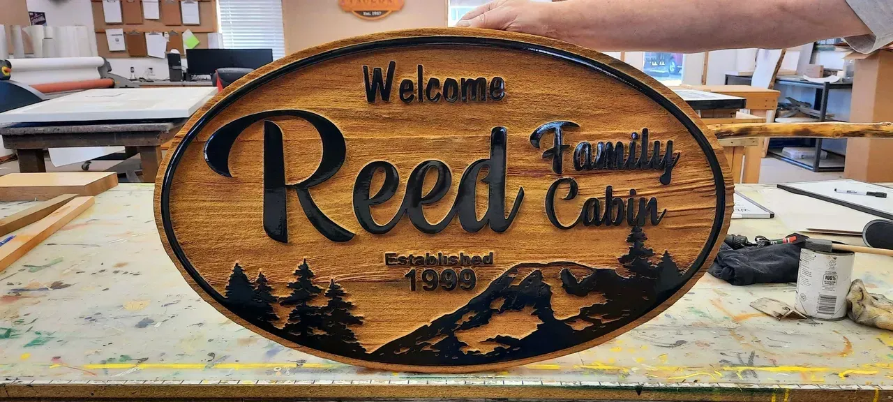 Oval wooden welcome sign reading “Reed Family Cabin” with pine tree and fish carvings created by Valley Sign