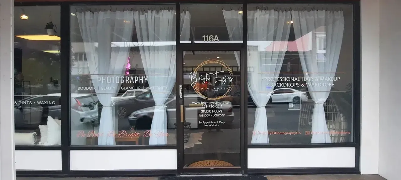 Storefront with black-framed windows, white curtains, and a glass door reflecting parked cars.