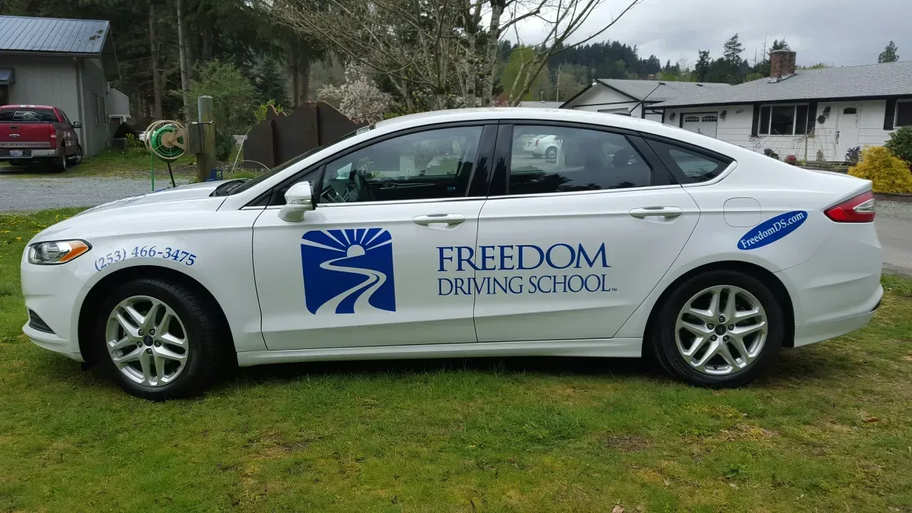 White sedan with “Freedom Living” logo parked on grass in a residential area