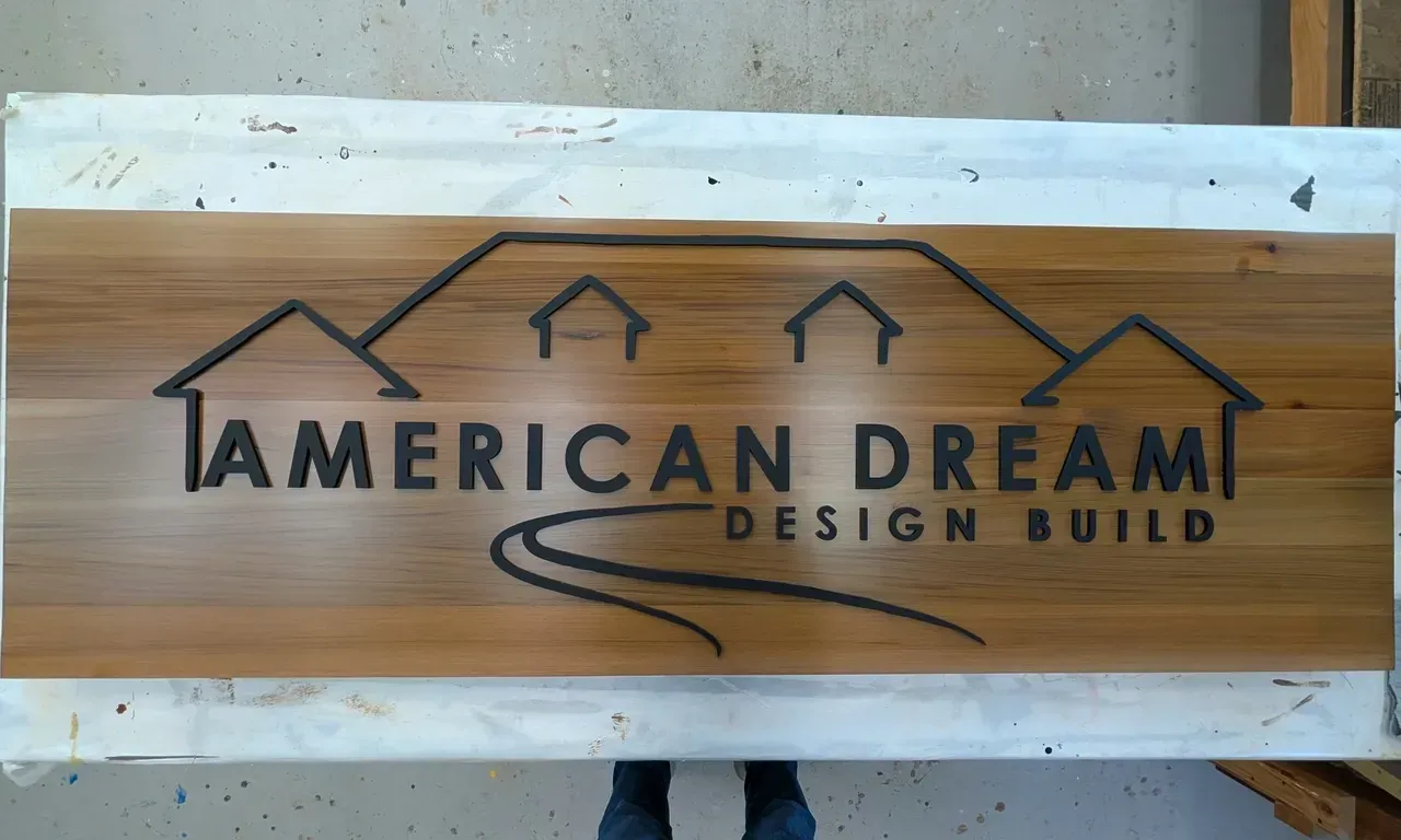 Wooden sign reading “American Dream Design Build” with house roof icons and a swoosh logo
