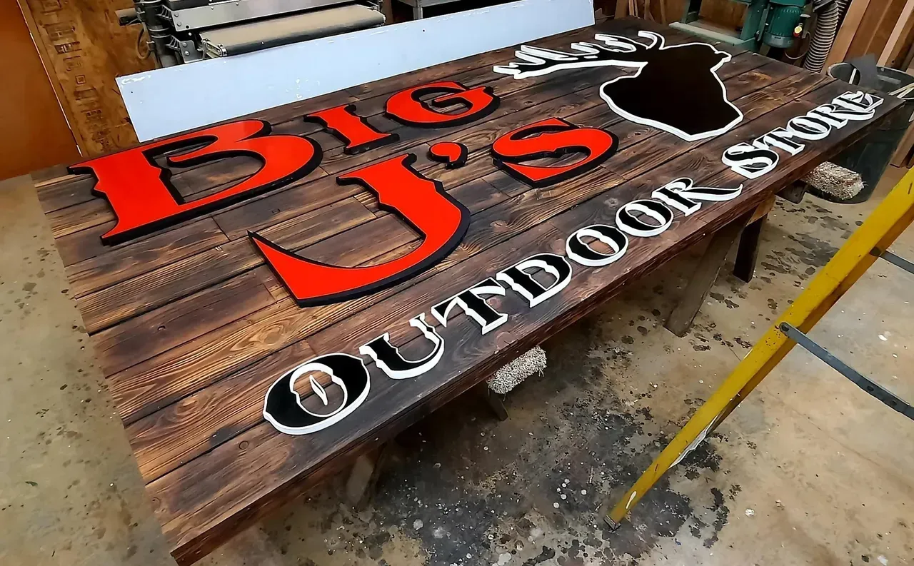 Wooden “Big J’s Outdoors” sign with red lettering, carved fish logo, and white script on a workbench.