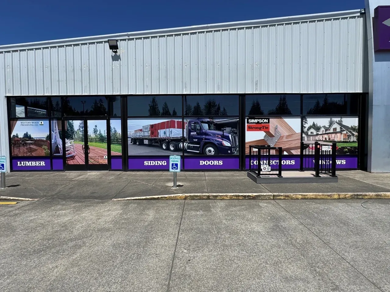Storefront with large windows, purple signage, and reflections under a gray metal facade on a sunny day