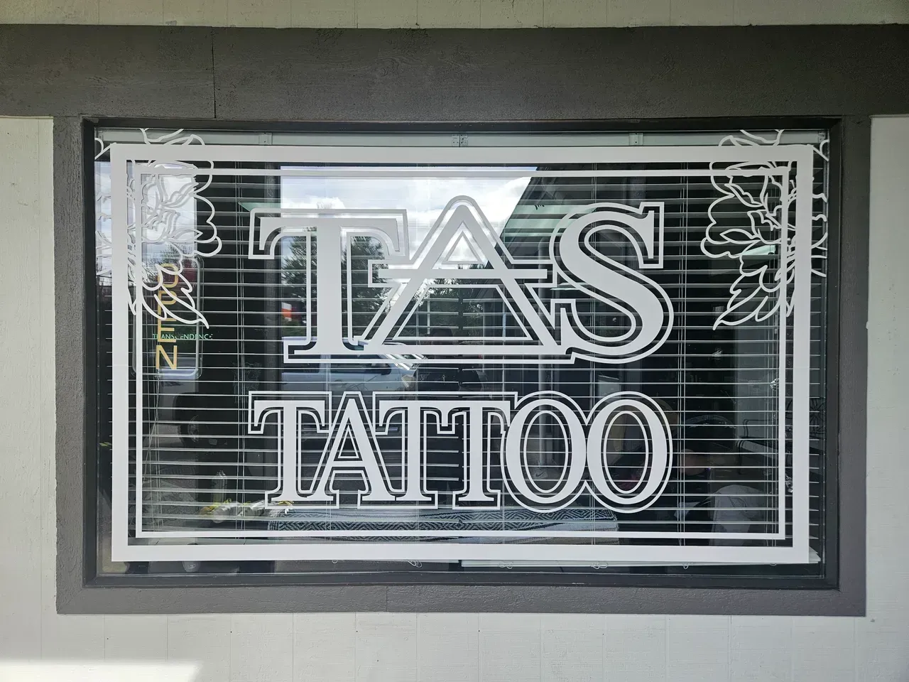 White neon “TAS TATTOO” sign in a storefront window with closed blinds