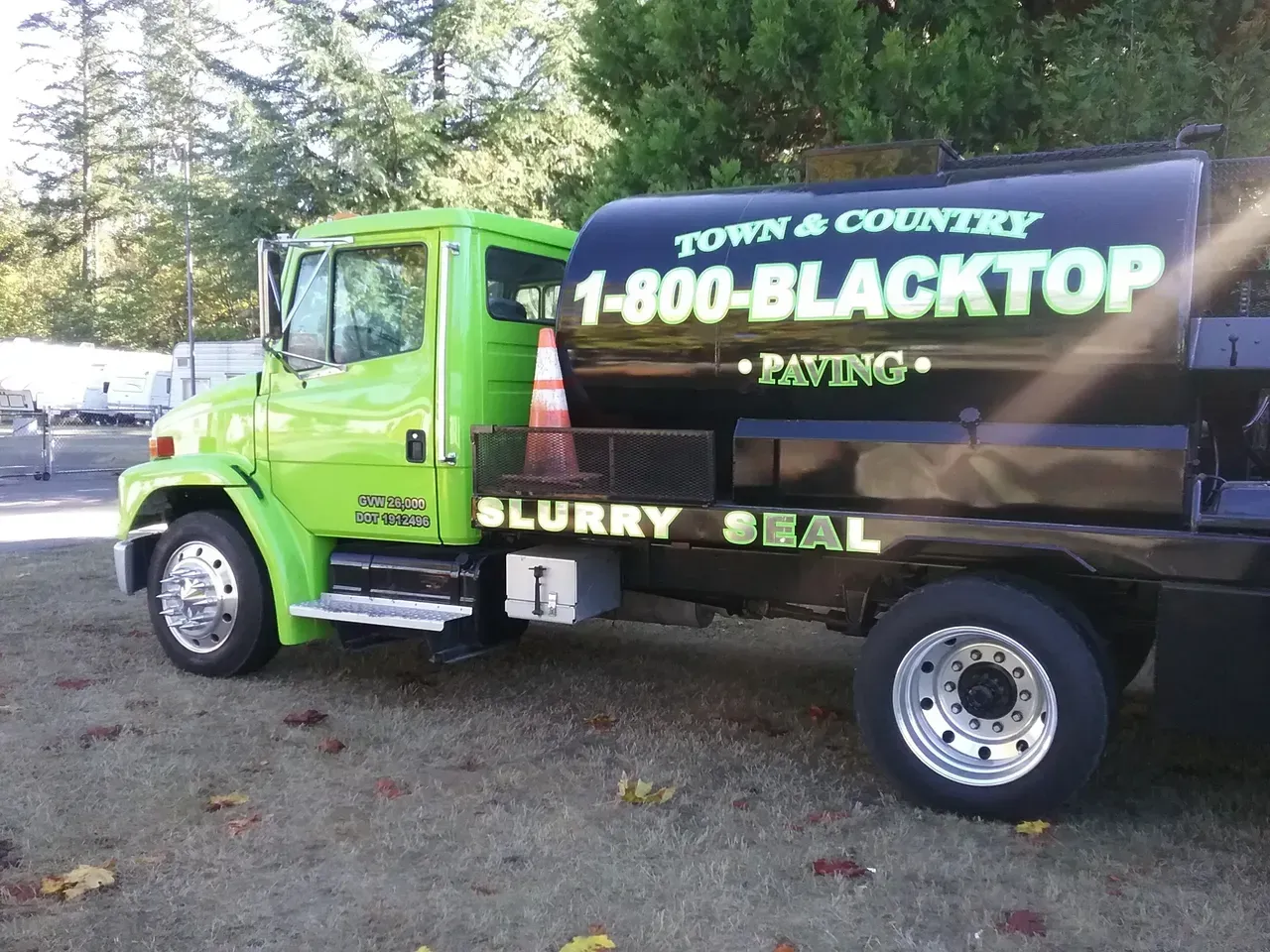 Green and black pavement truck with “1-800-BLACKTOP” branding parked outdoors near trees