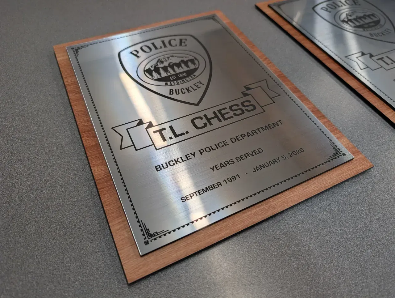 Plaques on a wall, including a silver police plaque with a wooden frame and “T.L. Chess” engraved.