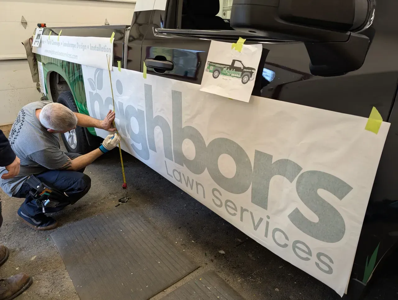 Worker applying a large vinyl banner to a vehicle in a shop, with tools and tape visible.