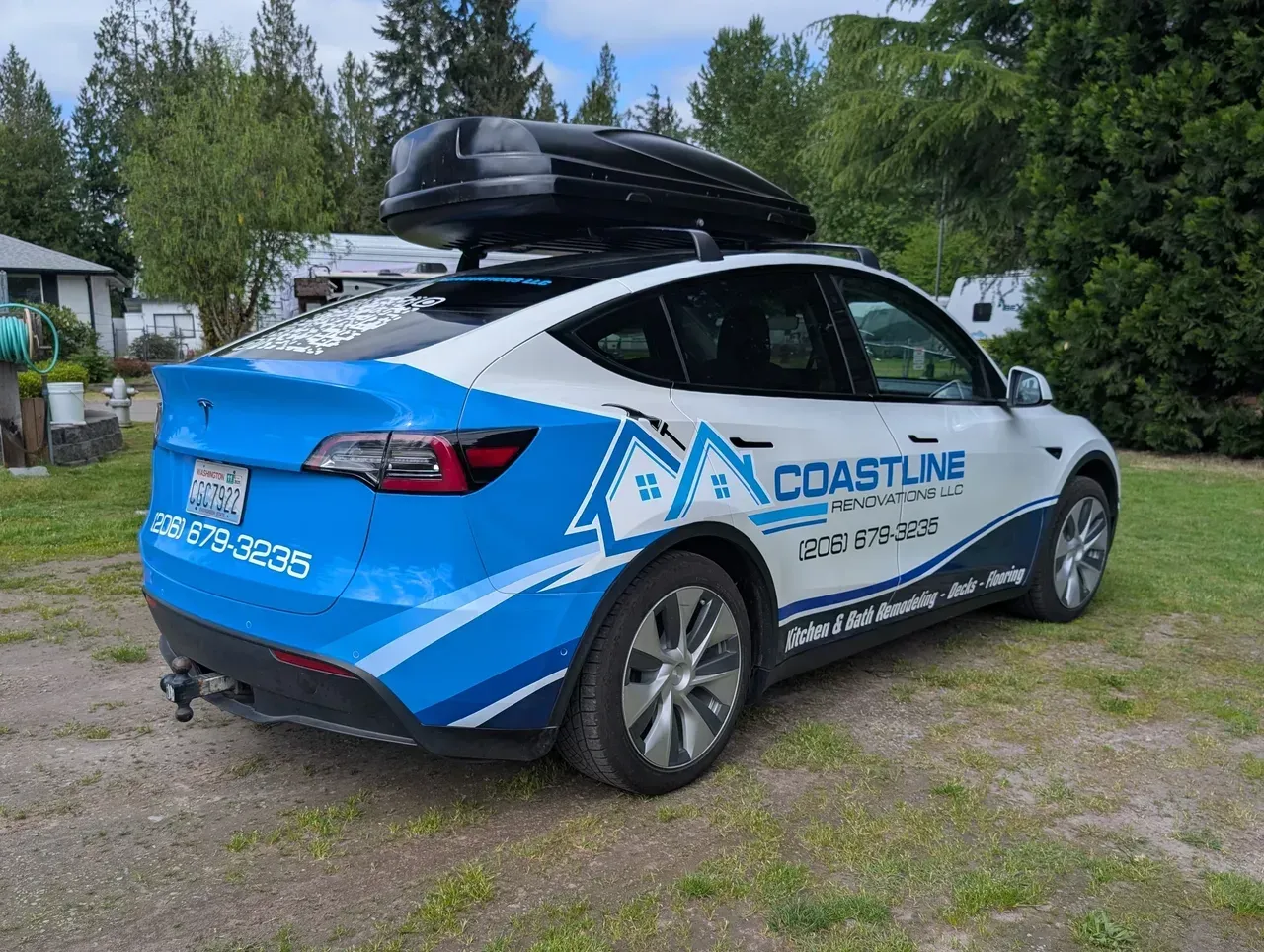 Blue and white Coasteer car with roof cargo box parked on a grassy area near trees and houses