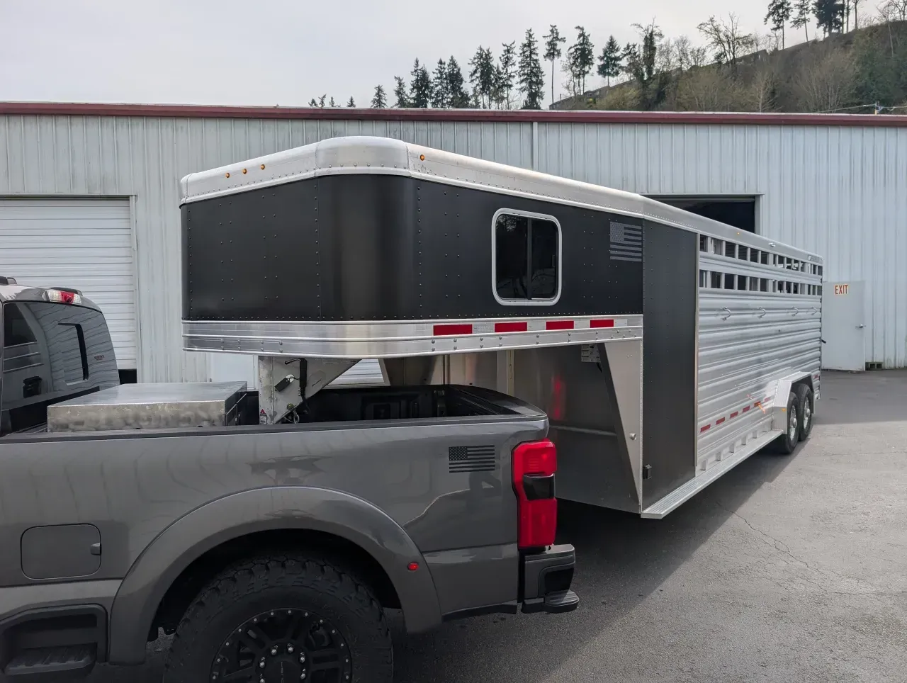 Gray pickup towing a silver livestock trailer parked outside a garage