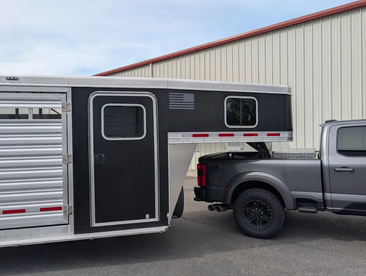 Silver pickup truck towing a black-and-silver truck camper beside a metal building.