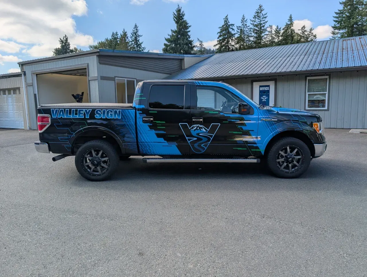 Blue and black pickup truck parked outside a building with trees in the background