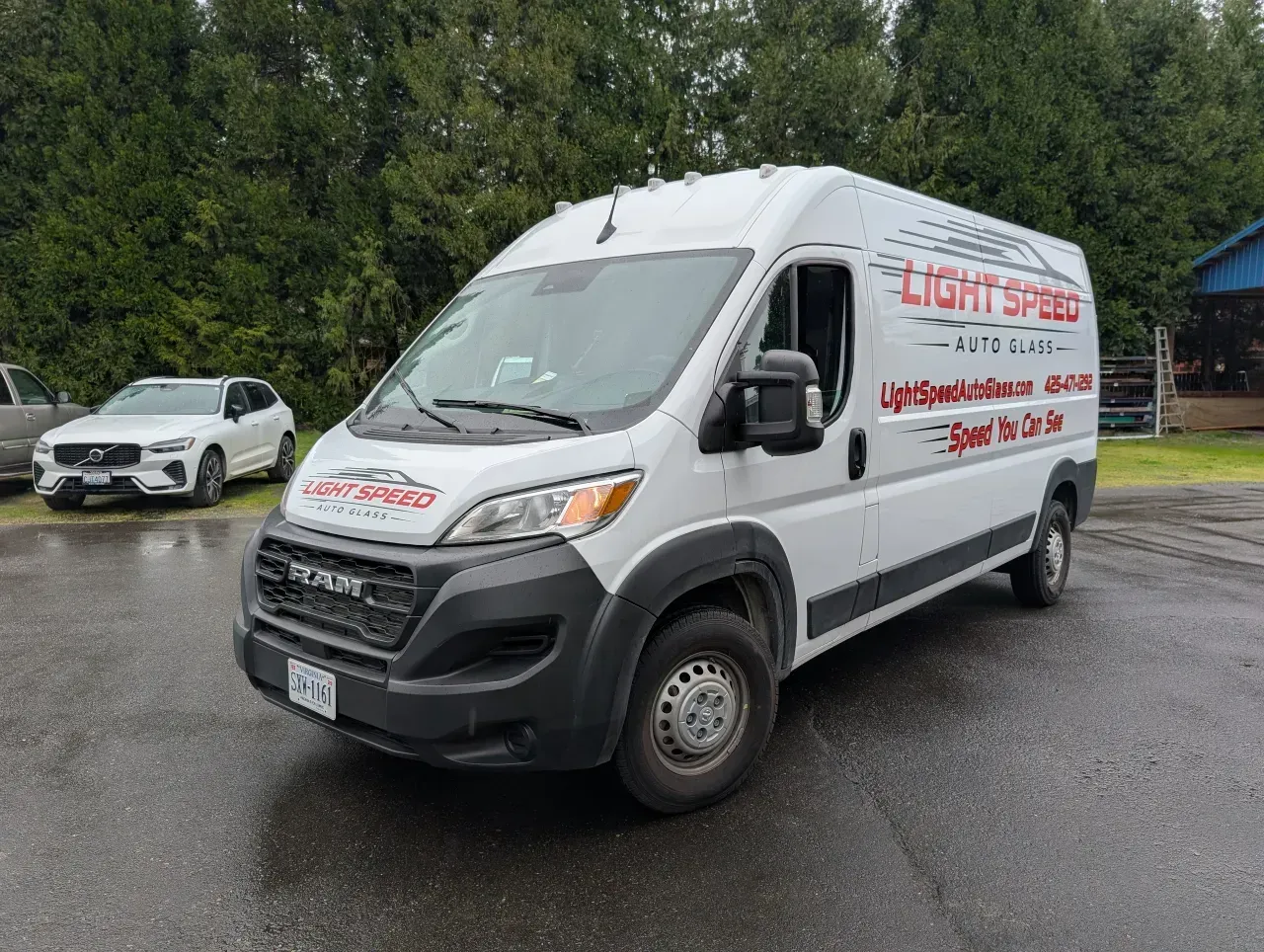 White cargo van with red lettering parked on a wet lot, with a silver car and trees in the background