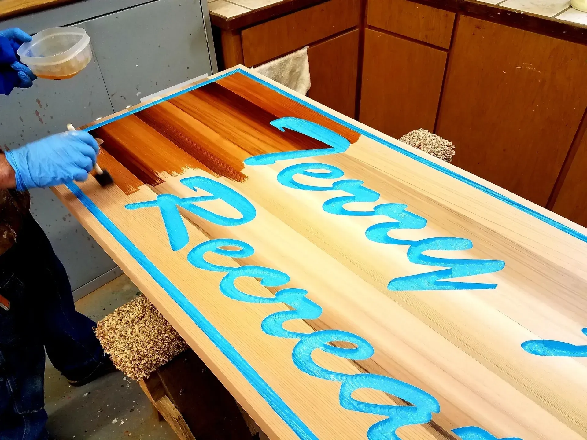 Person spray-painting blue script on a wooden sign in a workshop