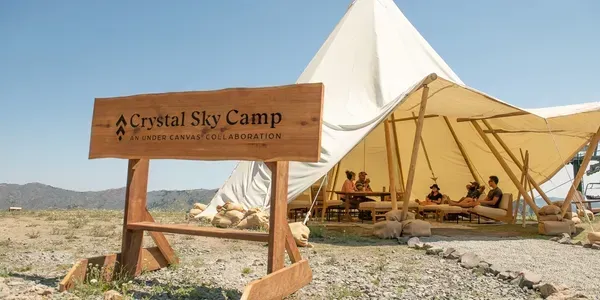 Desert camp entrance with “Crystal Sky Camp” sign, white tent, and seated guests under shade bunting.