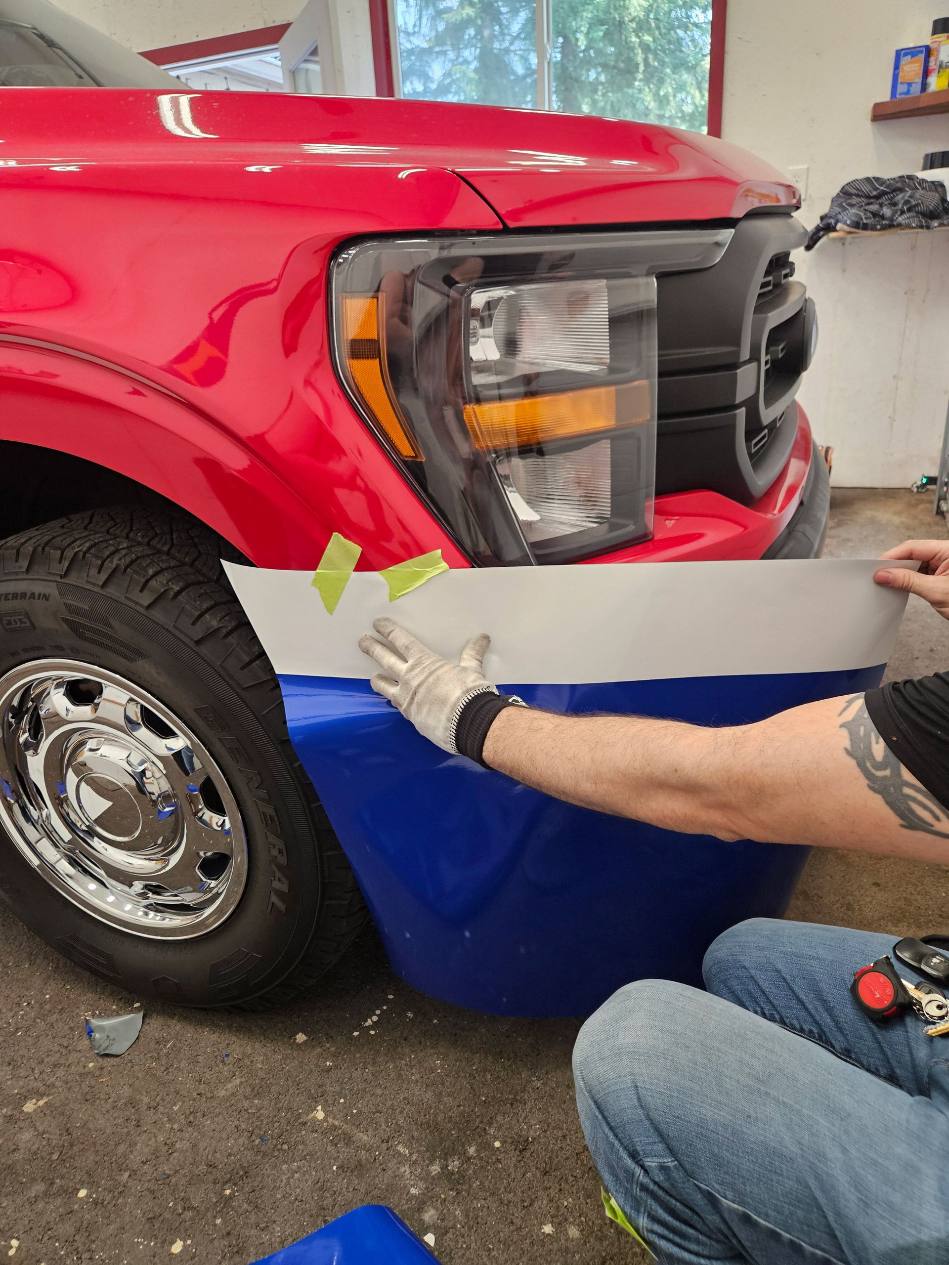 Person installing blue wrap on a red truck front bumper indoors