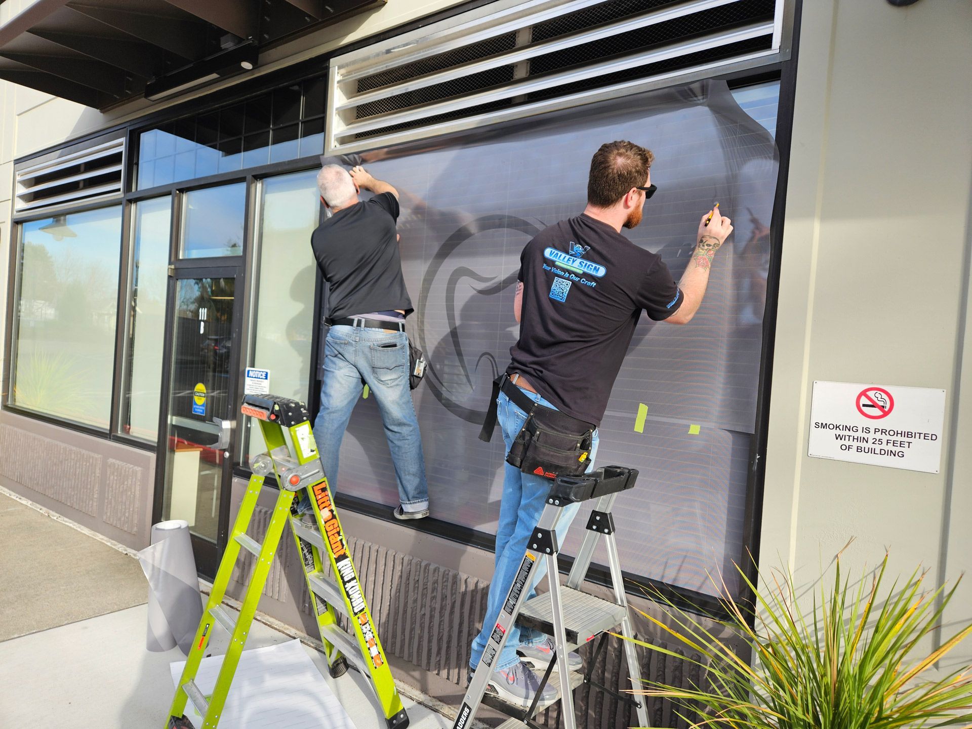 Two workers on ladders install a large window covering on a storefront exterior.