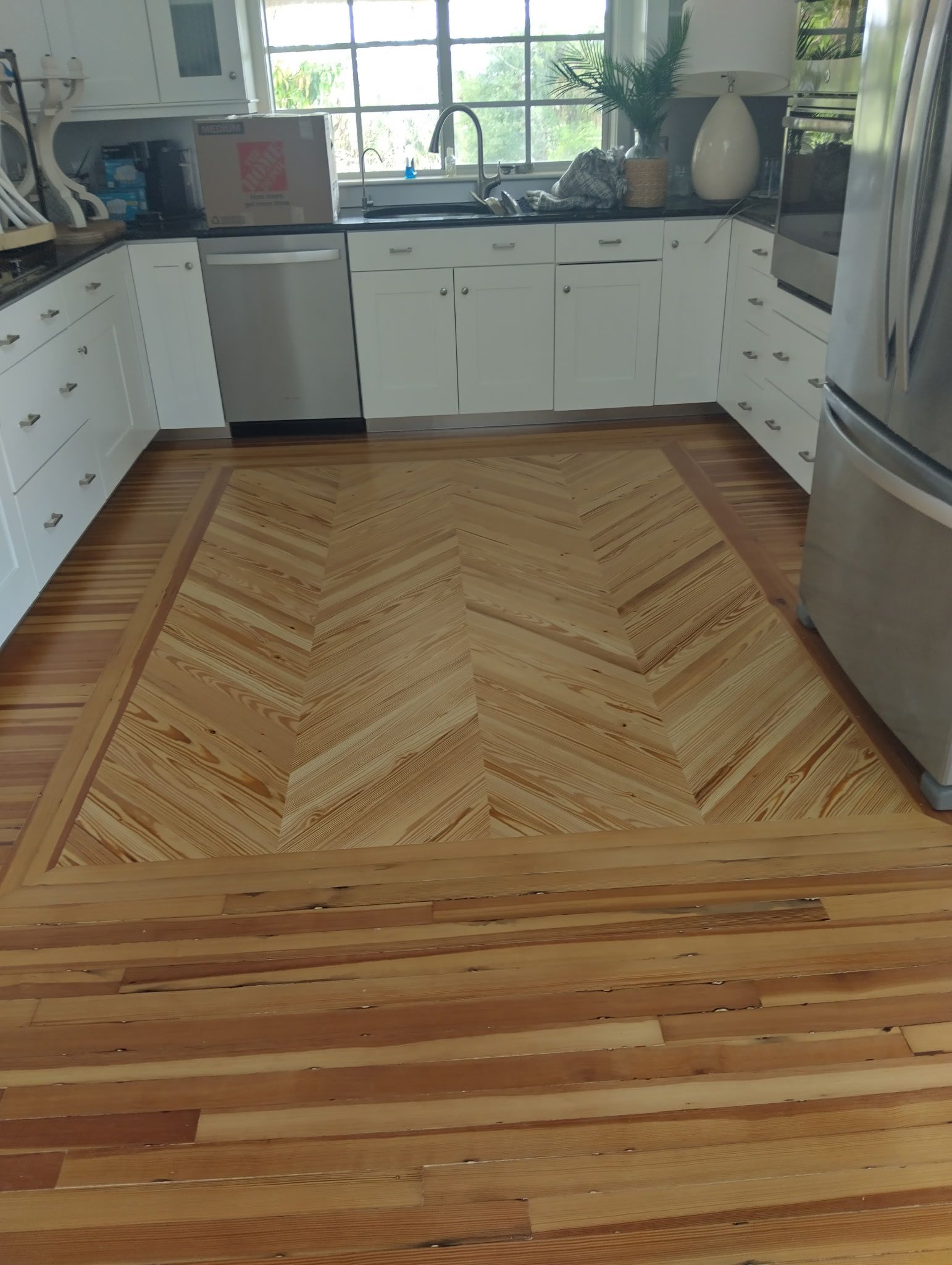 Bright kitchen with white cabinets, wooden floor, and a large woven rug by a sunlit window.
