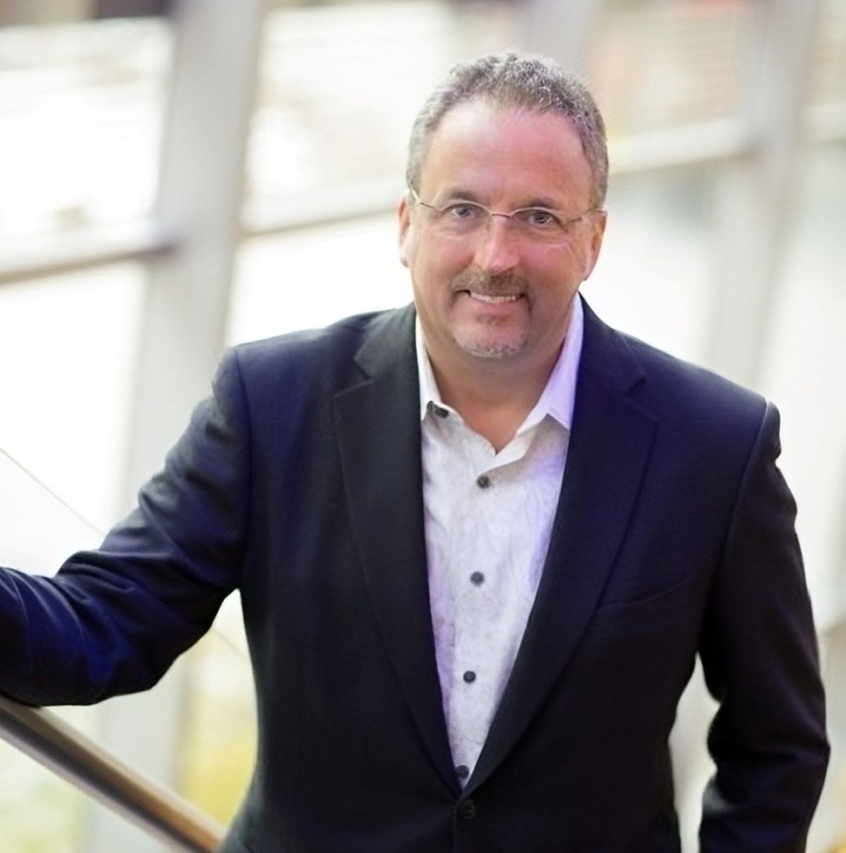 Man in a dark blazer and white shirt leaning on a railing, smiling in a bright indoor setting