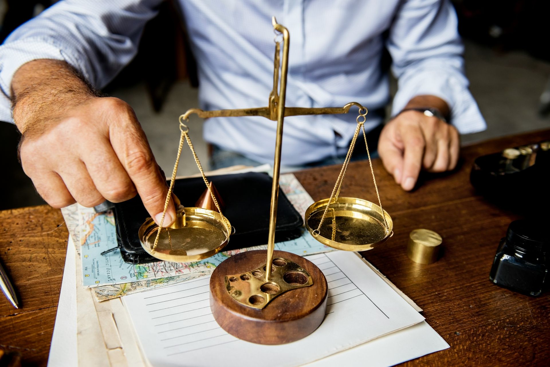 Hands examining a brass balance scale over papers on a wooden desk, with a law book and objects nearby