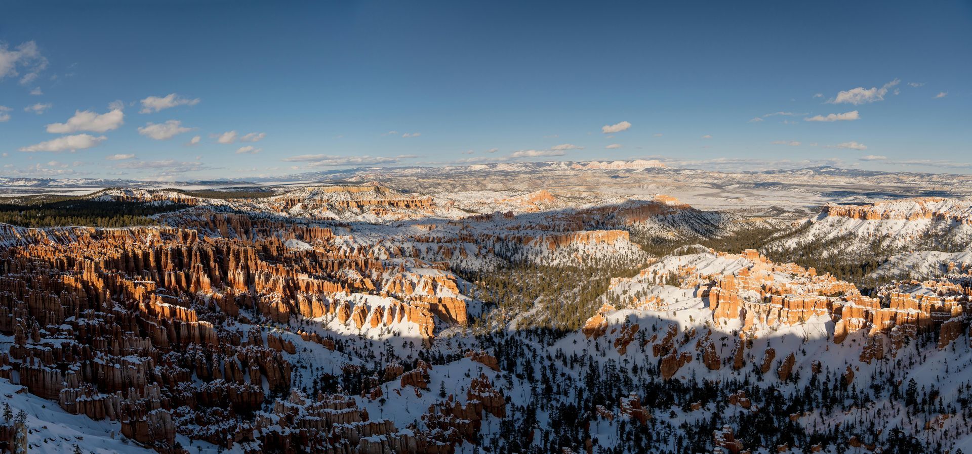 Snow-covered mountain valley at sunrise with rocky cliffs, pine forests, and a clear blue sky