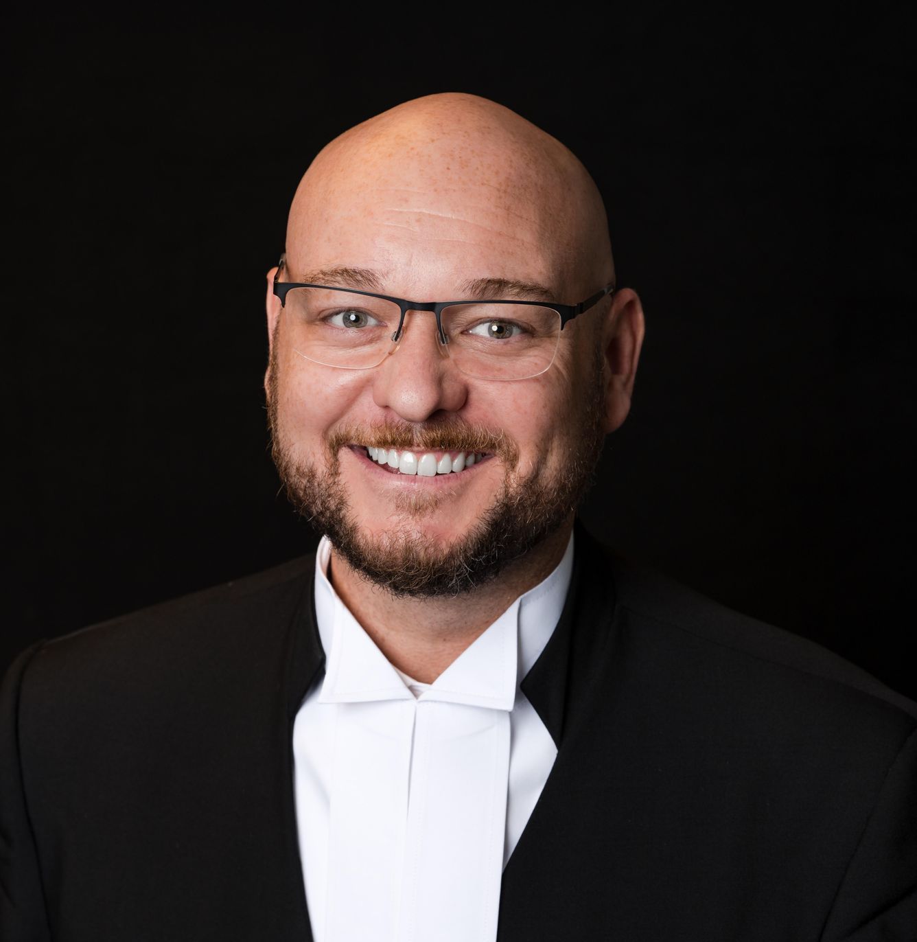 Bald man in glasses and suit smiling, likely a lawyer, against a black backdrop.