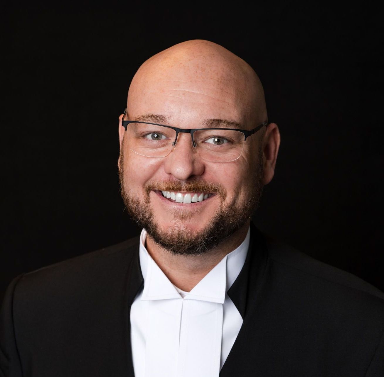 A bald man with a beard and glasses is wearing a tuxedo and bow tie.