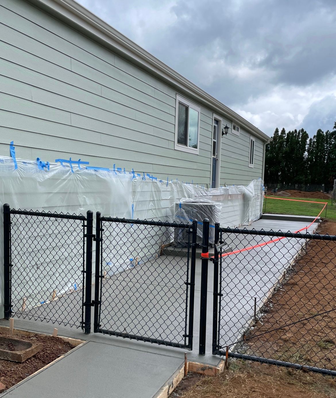A freshly poured concrete patio runs along a house covered in protective plastic sheeting, enclosed by a black chain-link gate.