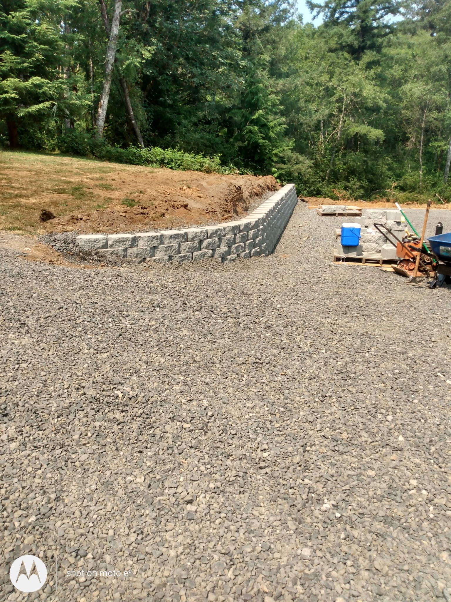 A curved stone retaining wall separates a gravel-covered yard from an elevated, grass-covered embankment in a forest.