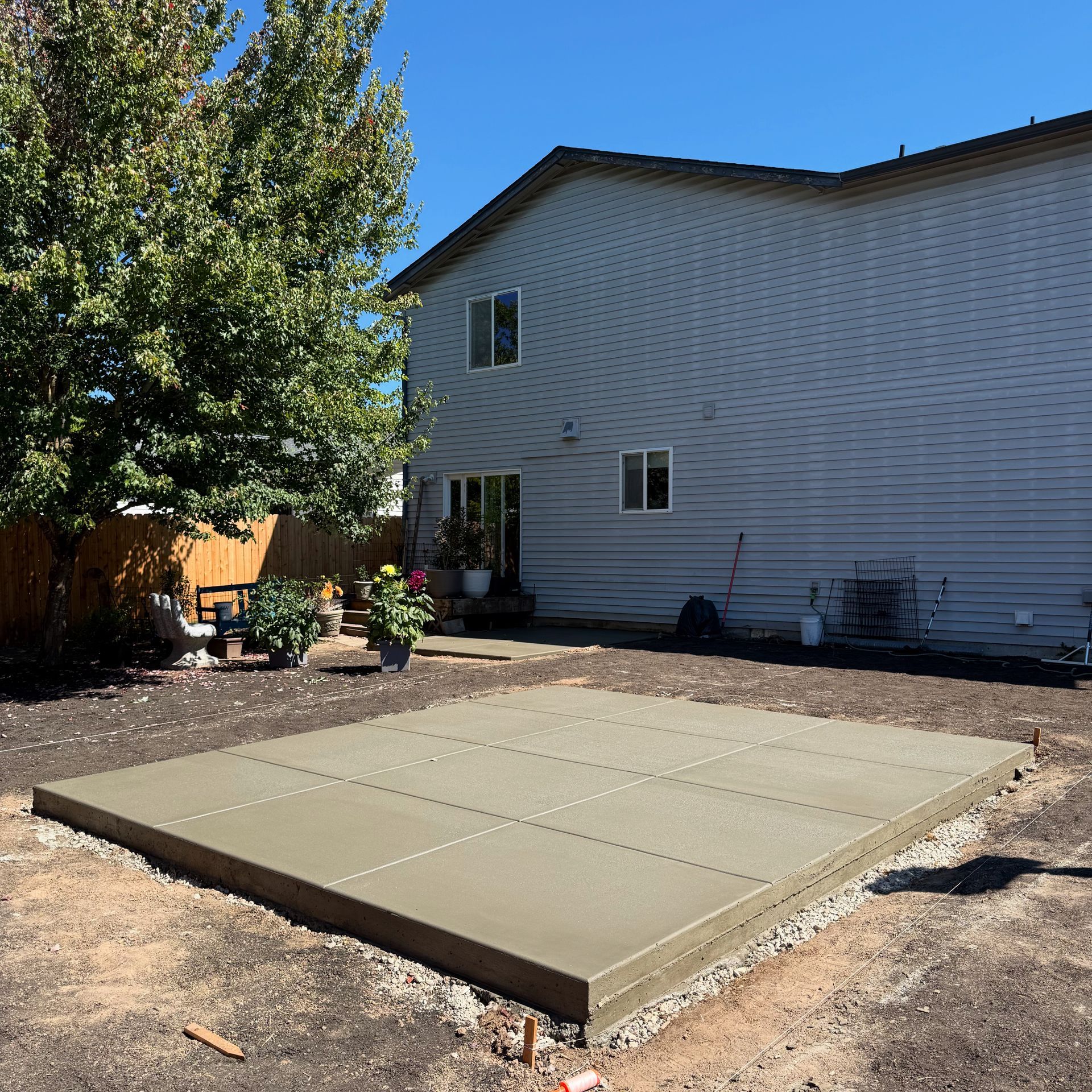 A freshly poured, square concrete patio slab sits in a dirt backyard next to a light gray two-story house.