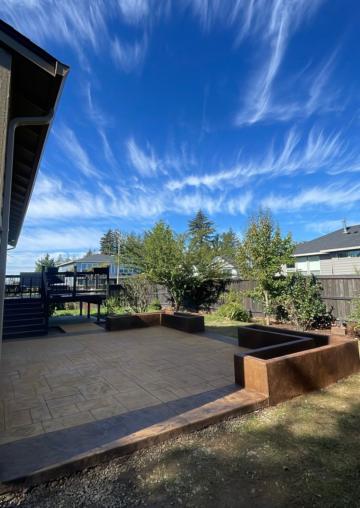 A brick patio with raised garden beds, a wooden deck, and trees under a bright blue sky with wispy clouds.