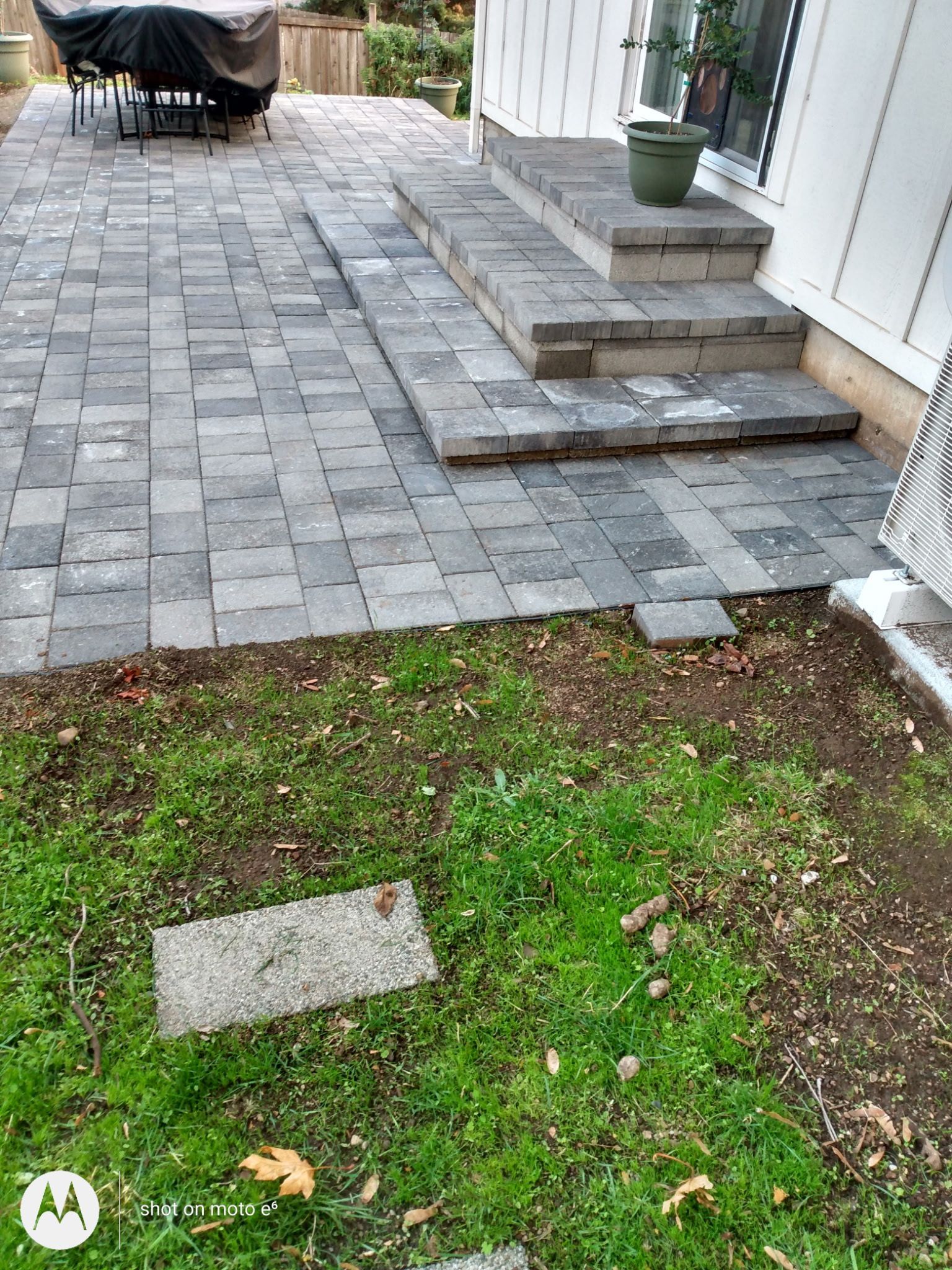 A gray paver patio leads to stone steps at a house entrance, with an area of dirt and sparse grass in the foreground.